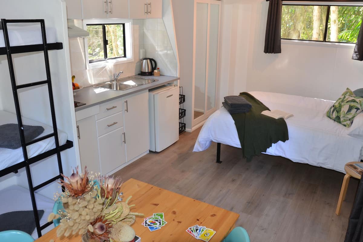 Una habitación compacta con kitchenette, una cama con manta verde, una mesa con cartas y una ventana con cortinas.