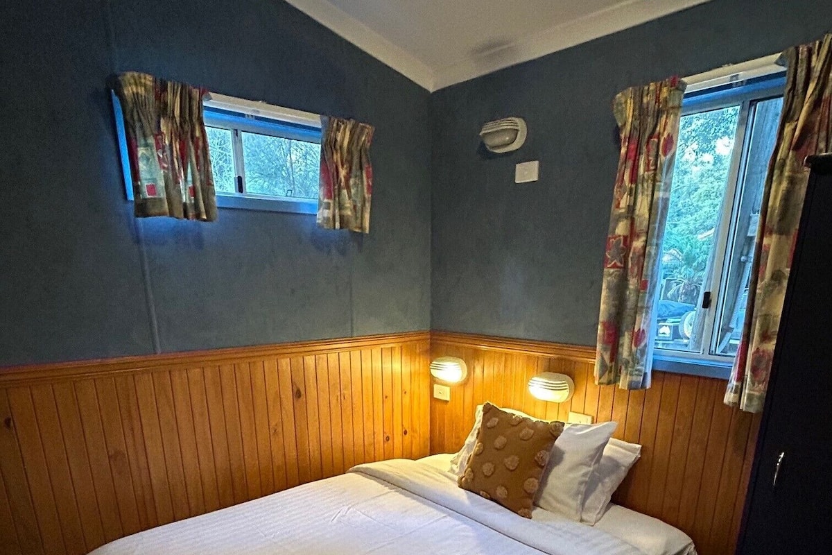 Una cama individual con una toalla doblada, paneles de madera en las paredes, dos ventanas con cortinas y una lámpara de noche.