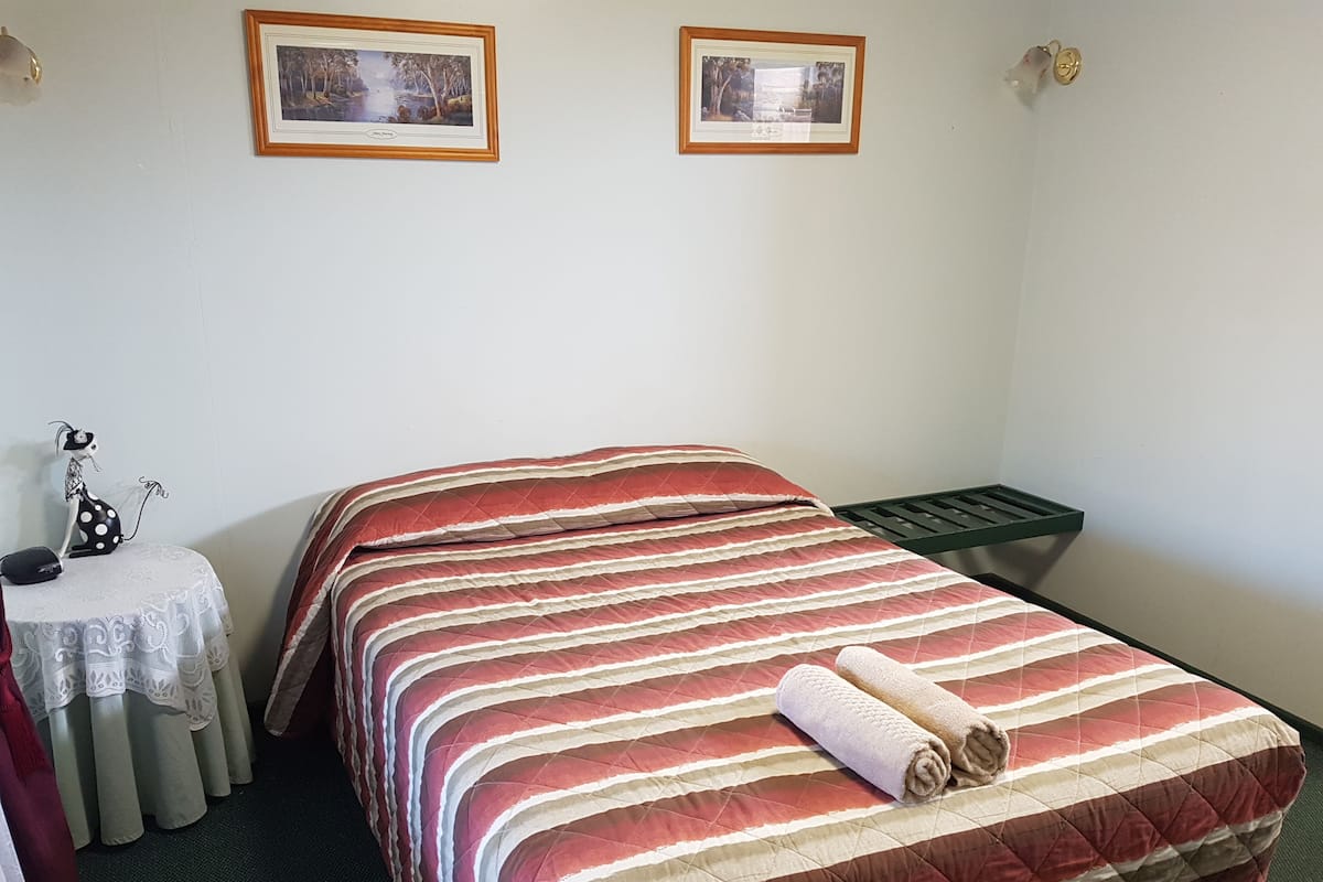 Ein Einzelbett mit einer gestreiften Bettdecke, ein kleiner Tisch mit weißer Tischdecke und zwei gerahmte Bilder an der Wand.