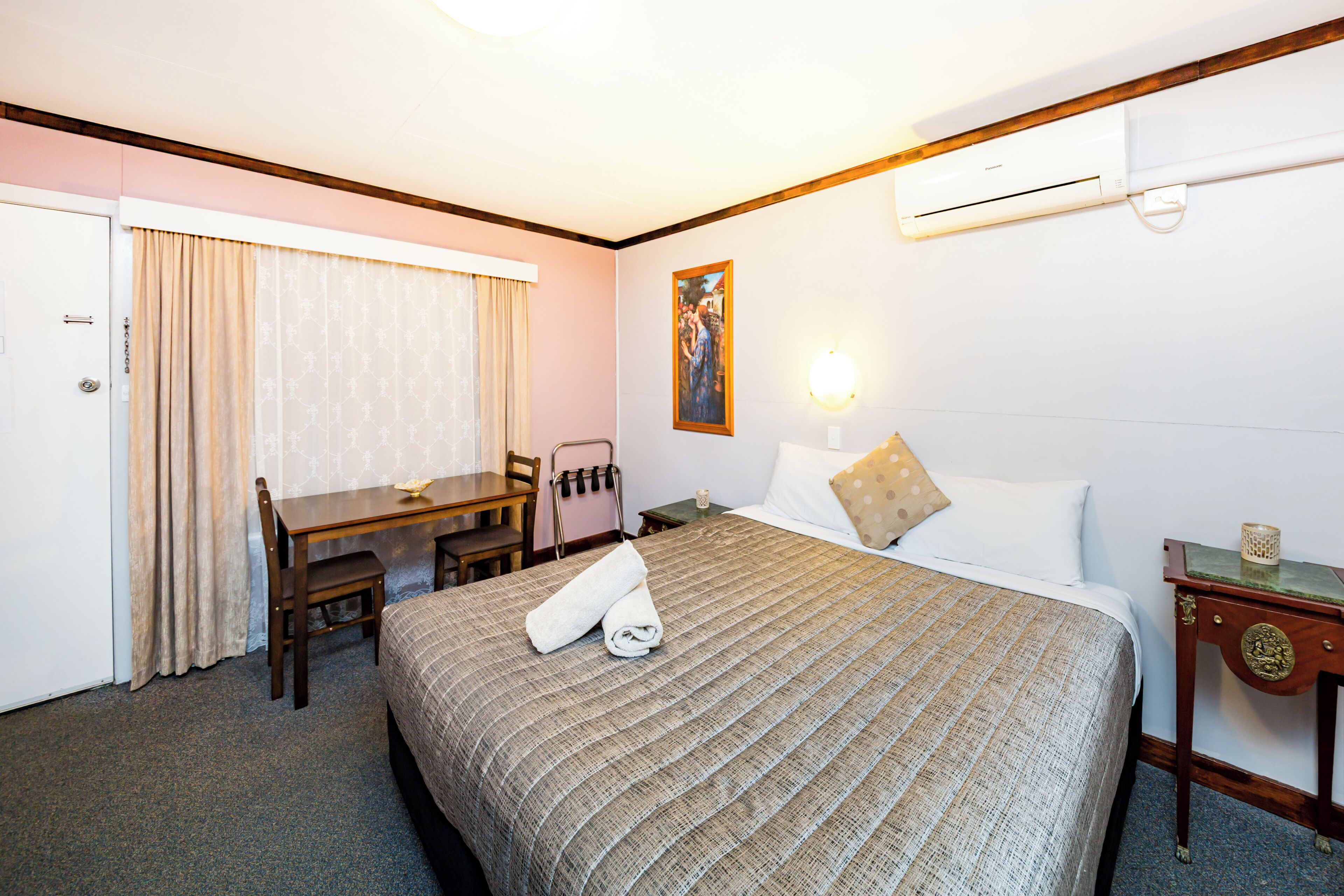 A hotel room with a bed, a desk, a chair, a table, a painting on the wall, and an air conditioning unit.