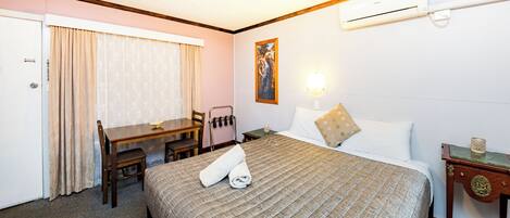 A hotel room with a bed, a desk, a chair, a table, a painting on the wall, and an air conditioning unit.