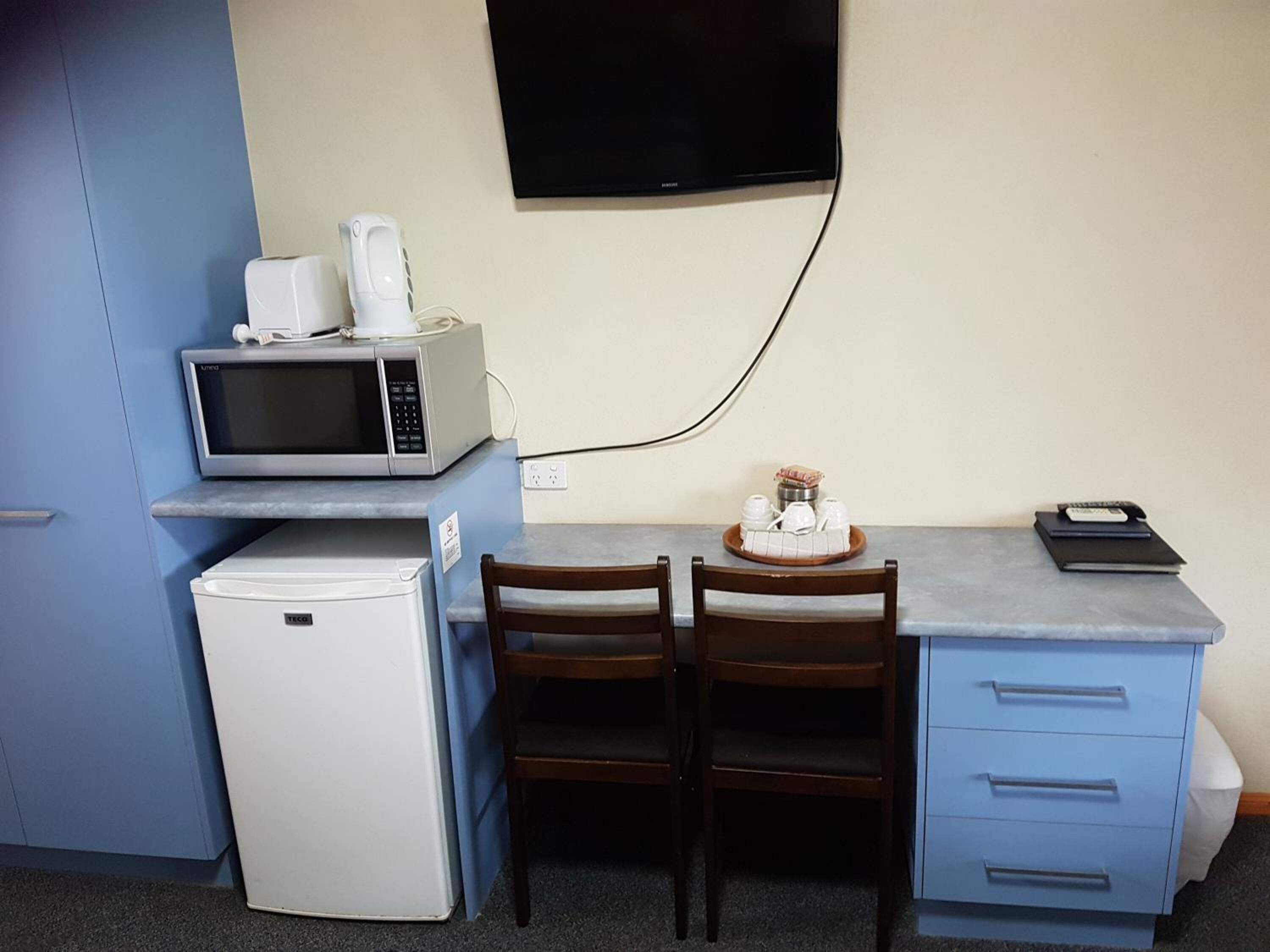 A compact room with a kitchenette, a microwave, a mini-fridge, a small table with chairs, a TV mounted on the wall, and a blue cabinet with drawers.