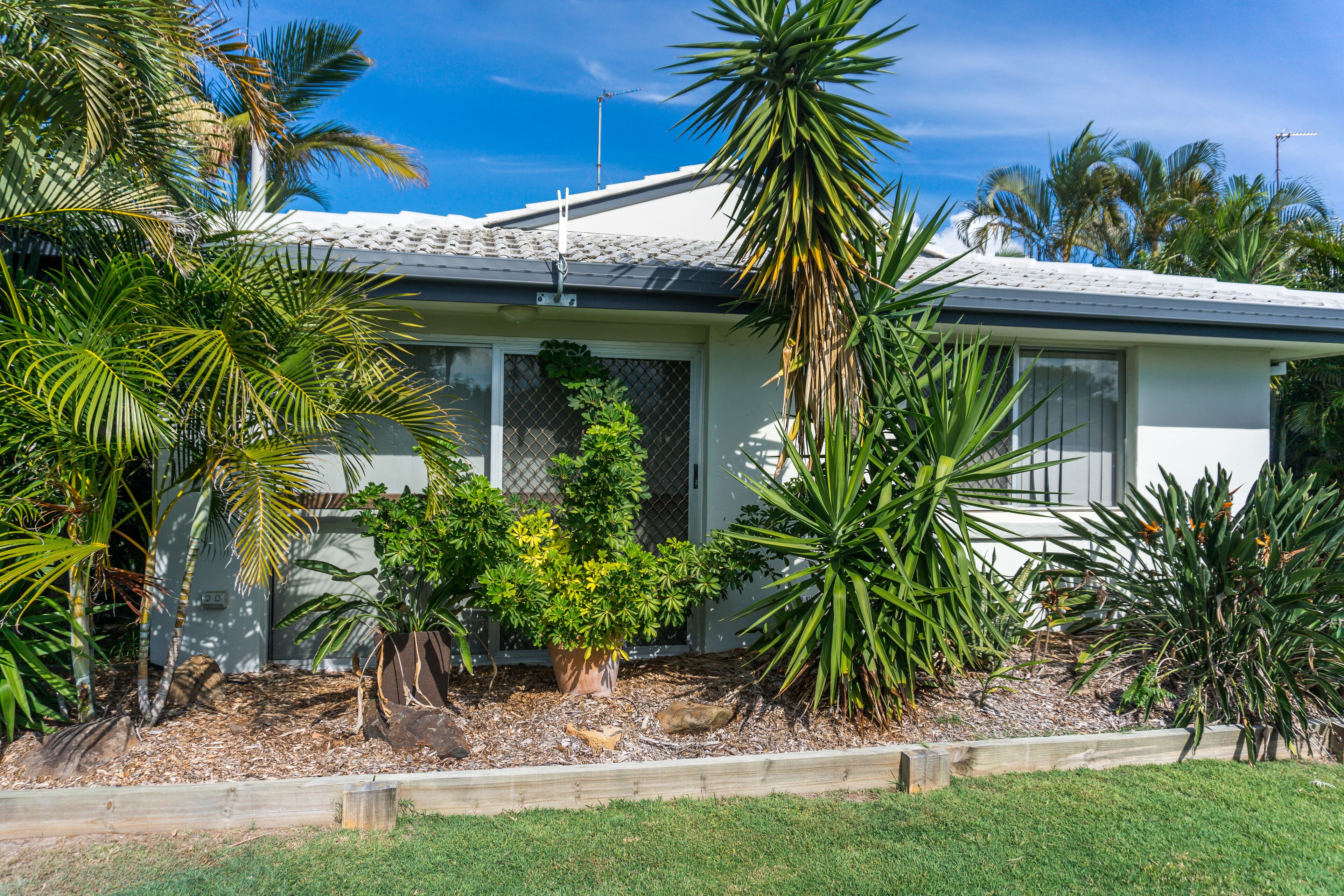 Une maison avec un toit en pente, entourée de diverses plantes tropicales et d’une pelouse bien entretenue.