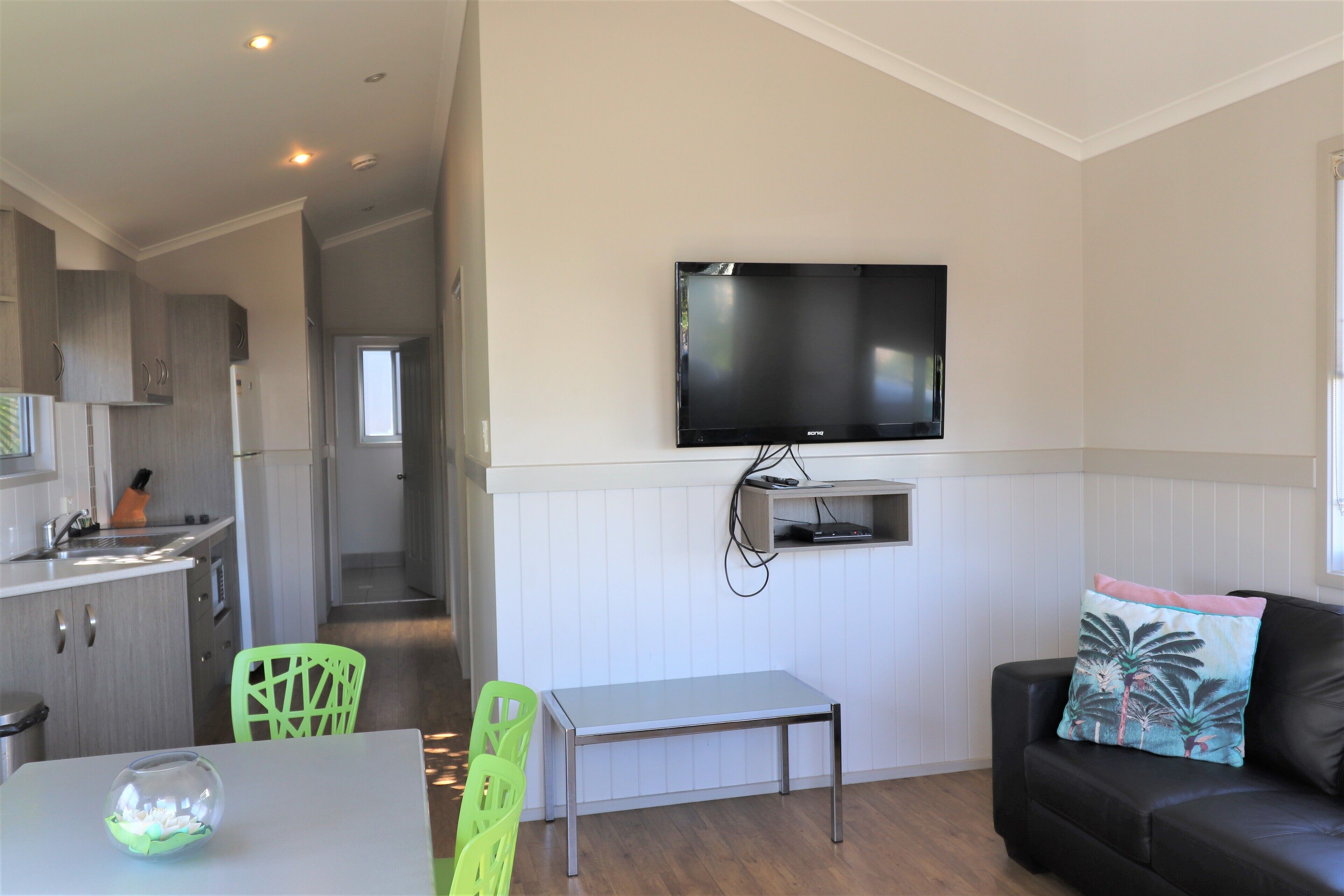 Un espacio habitable compacto con kitchenette, un área de comedor con mesa y sillas, un sofá de cuero negro con un cojín decorativo y un televisor de pantalla plana montado en la pared.