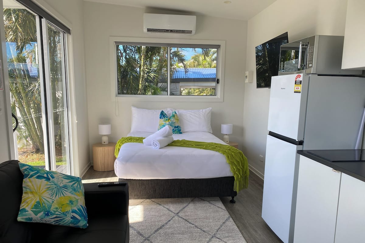 A hotel room with a bed, a sofa, a kitchen area, and a window with a view of palm trees.