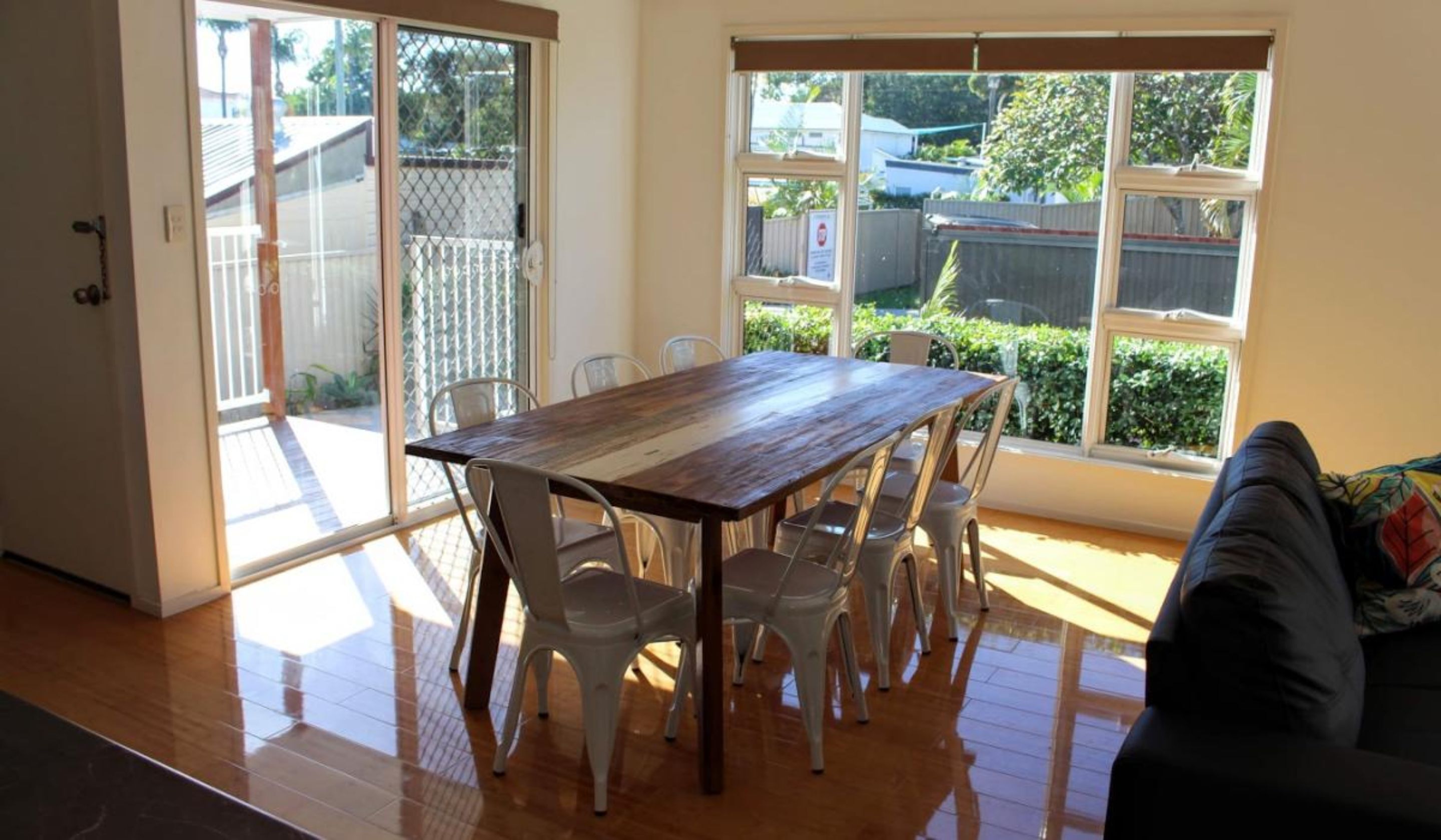 A dining area with a wooden table and white chairs, large windows with a view of a backyard, and a sliding glass door leading to an outdoor space.