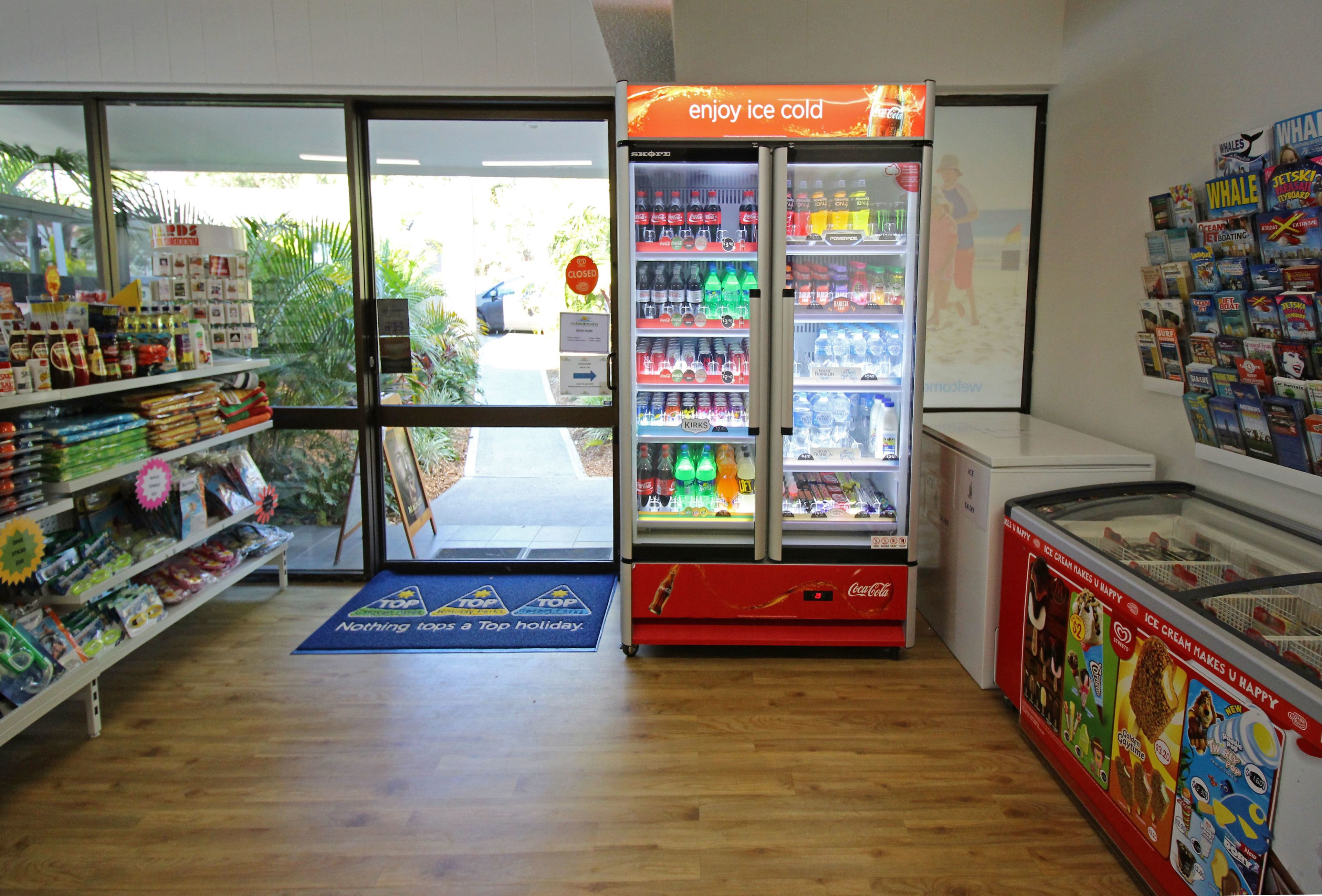 Interior de una tienda con un refrigerador de Coca-Cola, estantes llenos de diversos productos y una exhibición de helados.