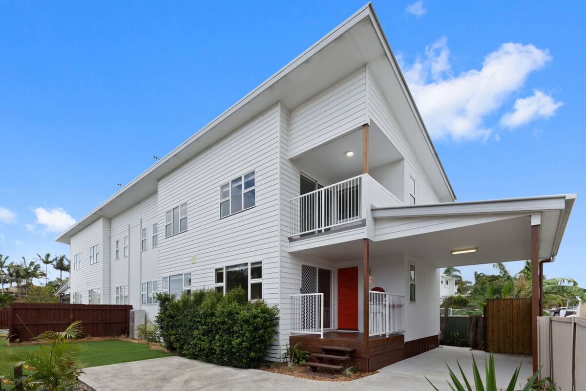 A two-story residential building with a balcony on the second floor, a front porch, and a red door.