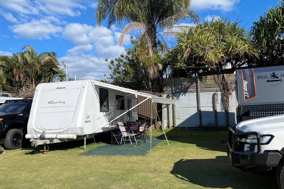 A Jayco Sterling RV with a covered patio area, parked in a grassy yard with palm trees and a corrugated metal building in the background.