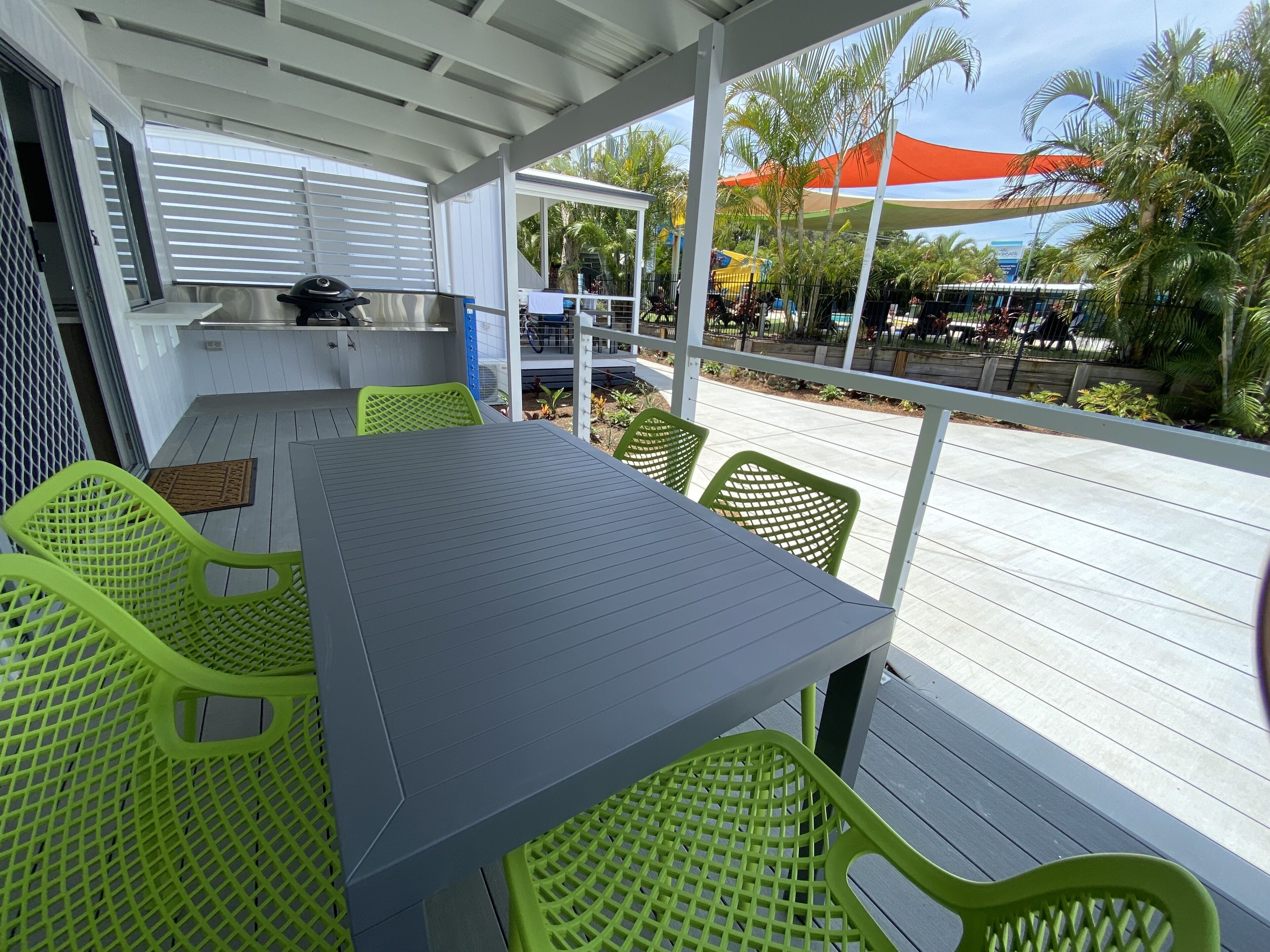 Un espace extérieur abrité, aménagé pour les repas, avec une table et des chaises, un barbecue et une vue sur la piscine et les palmiers.