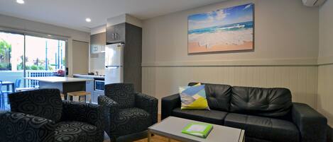 A modern living room with a black leather sofa, patterned armchairs, a glass coffee table, and a painting of a beach scene on the wall.