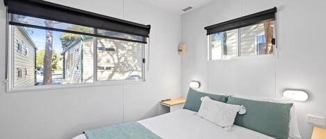 A bedroom with a bed, two bedside tables, a nightstand, a mirror, and a window with a view of buildings and trees.