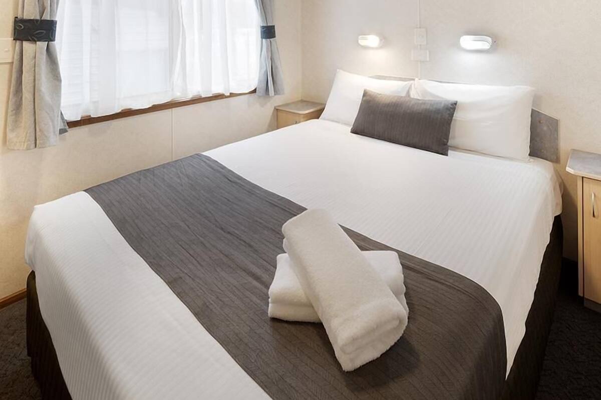 A neatly made bed with white and gray linens, a folded white towel, and a nightstand with a lamp.
