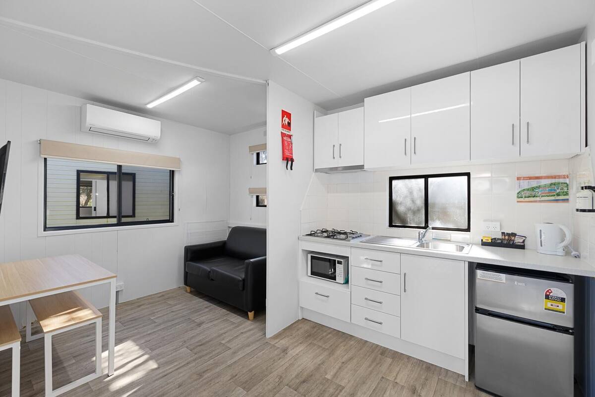A modern kitchen with white cabinets, a black sofa, a dining table, and a window with blinds.