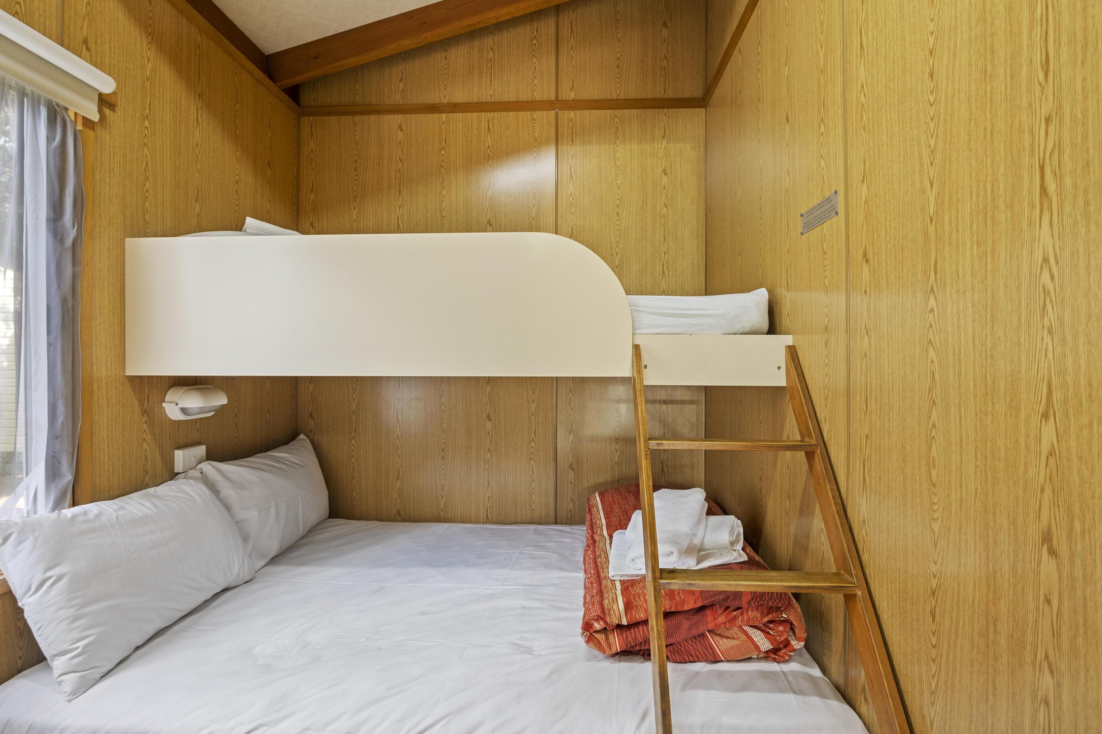 A bunk bed with white bedding, a wooden ladder, and a small shelf with towels.