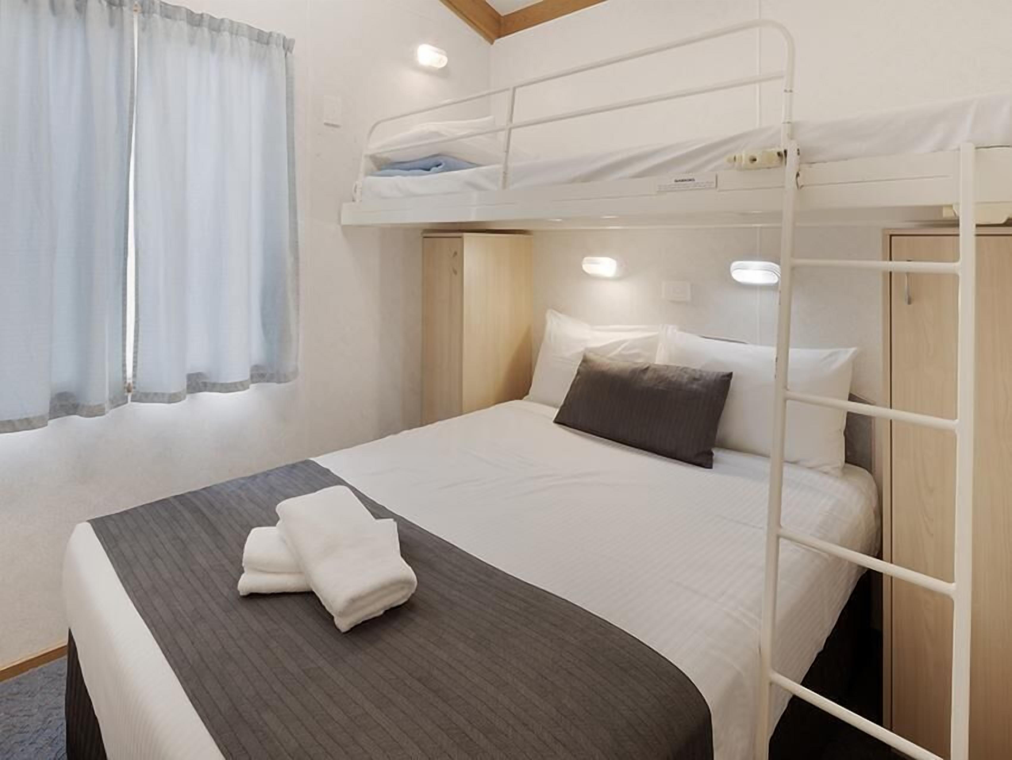 Habitación con literas, una cama bien hecha, una toalla doblada y una ventana con cortinas.