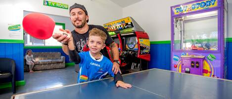 Une table de ping-pong dans une pièce où l’on peut voir une pancarte indiquant « salle de télévision », une borne d’arcade sur le thème de Toy Story et un canapé.