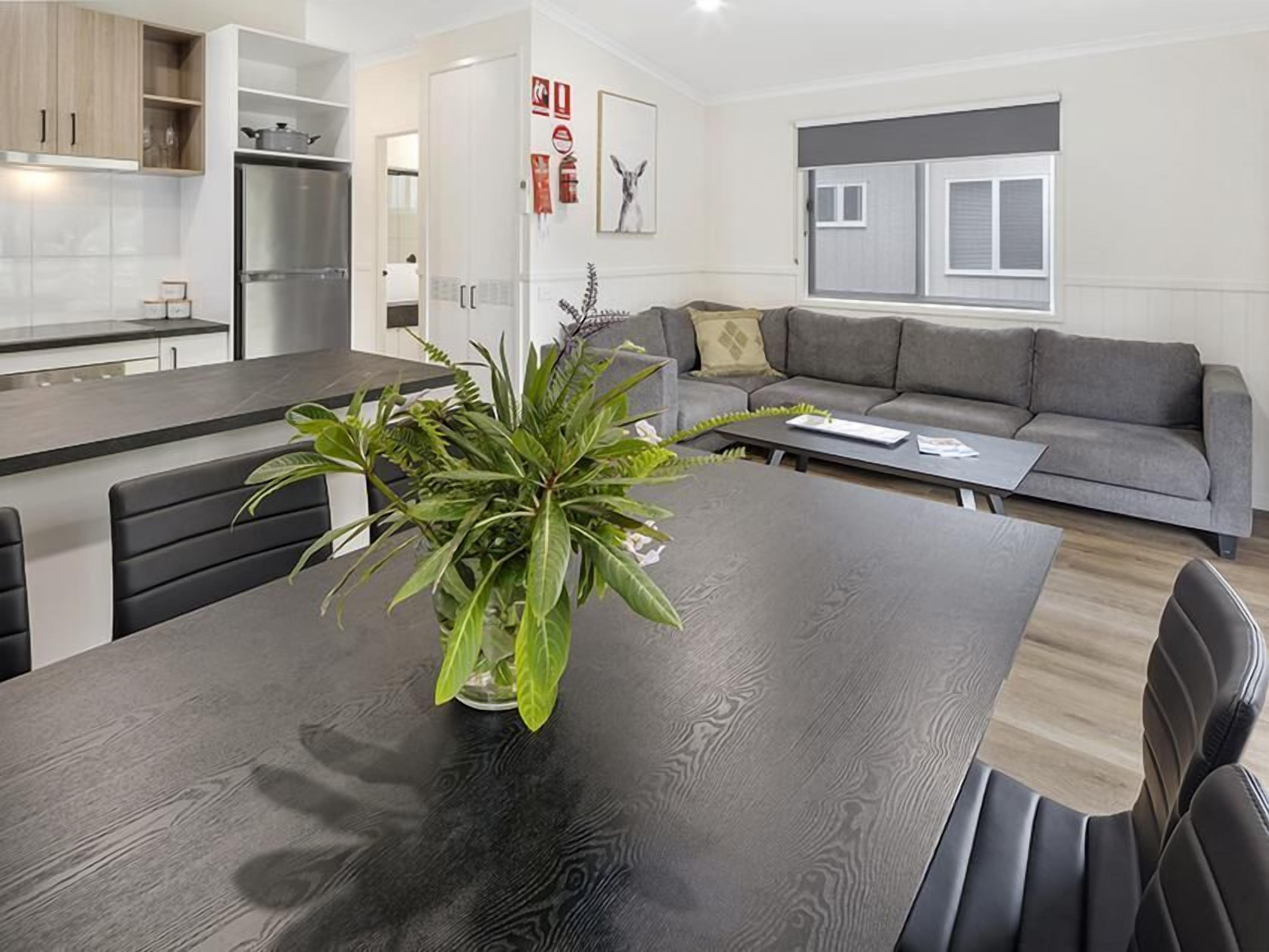 A modern kitchen with a central island, a dining table with chairs, a grey sofa, and a large window.