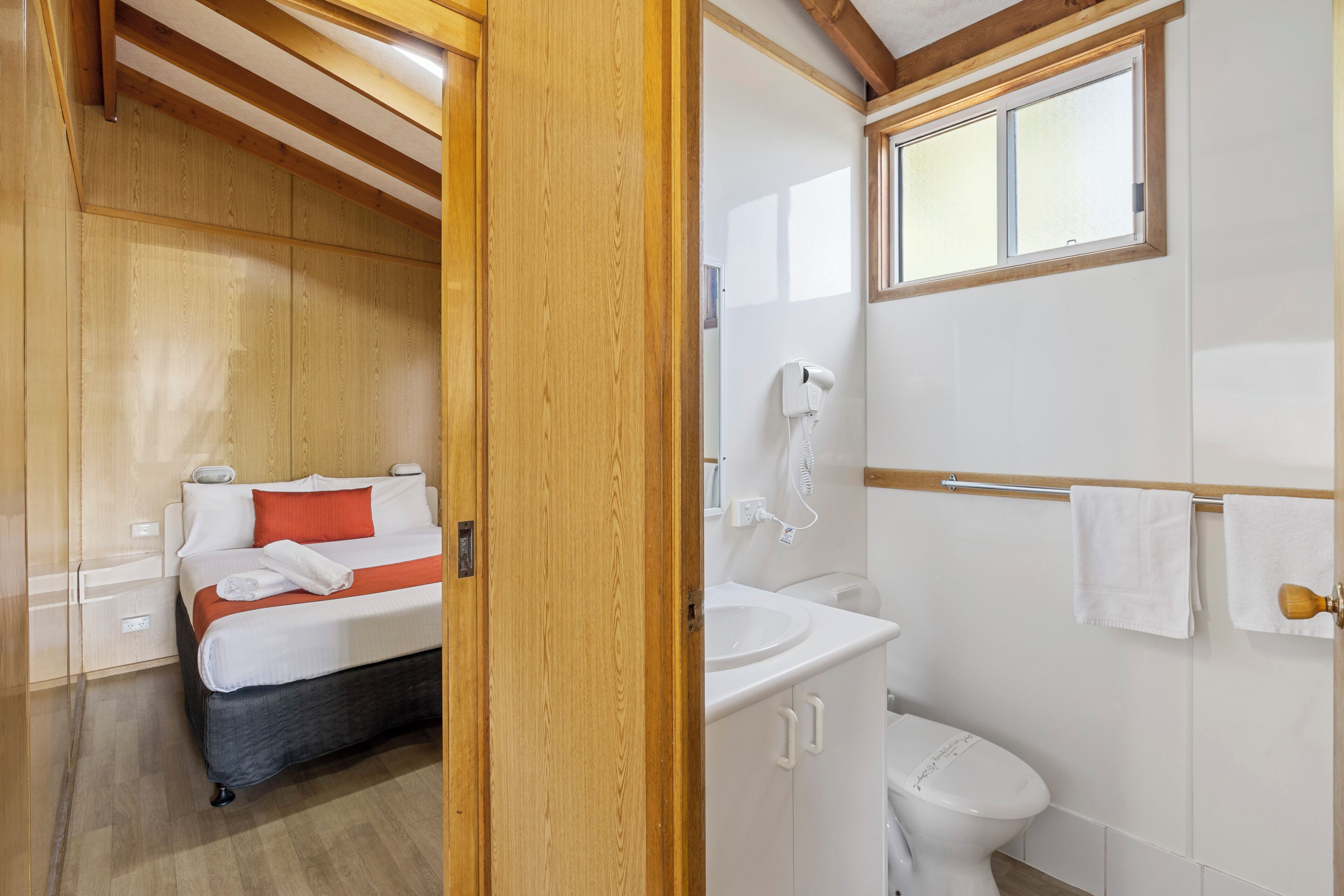 A bedroom with a bed, a bedside table, and a wooden ceiling. The room has a window and a door.