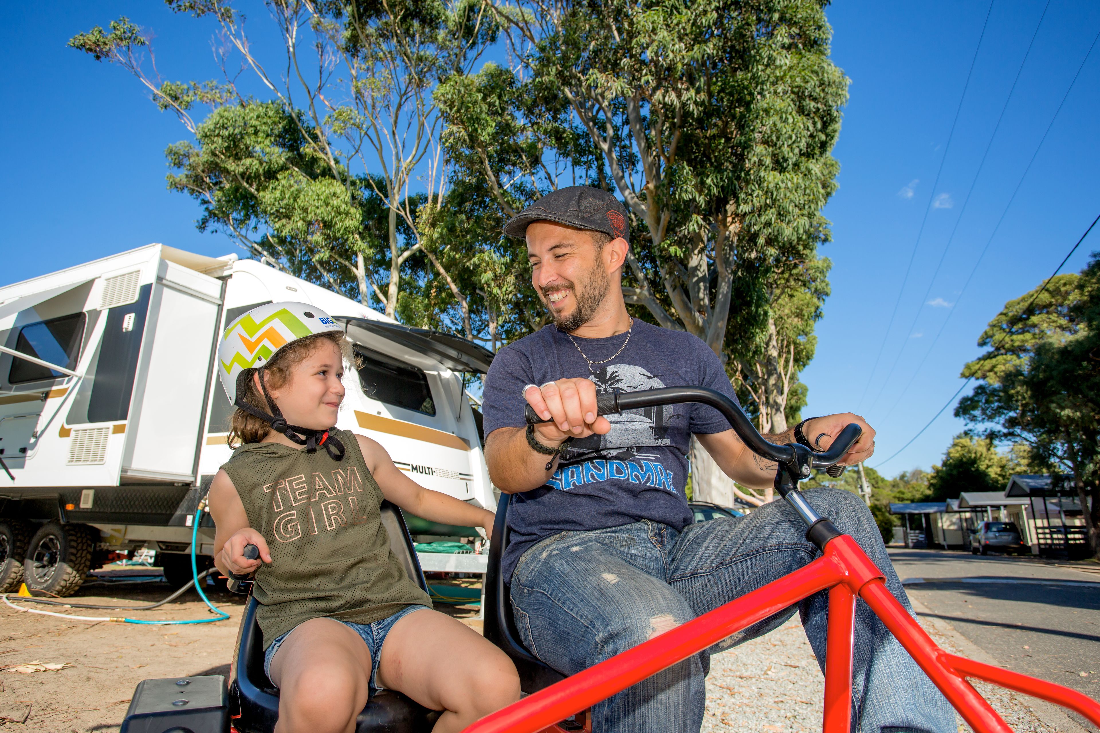 A person riding a bicycle with a red frame, another person sitting beside them, and a white RV in the background.