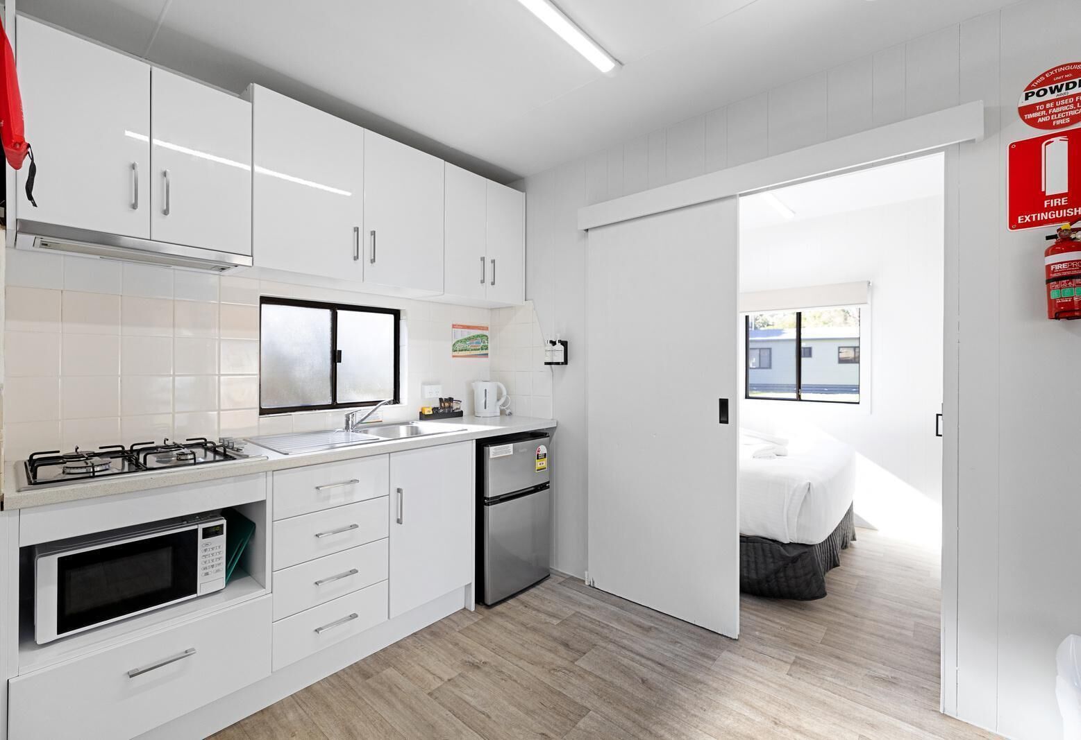 A modern kitchen with white cabinets, a gas stove, a microwave, and a dishwasher. There is a window above the sink and a door leading to a bedroom.
