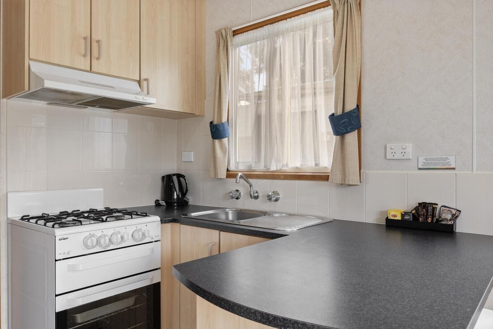 Cocina con estufa a gas, horno y lavabo. Se ve una ventana con cortinas de gasa y un estante pequeño con utensilios de cocina.