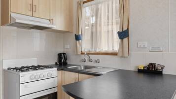 Cocina con estufa a gas, horno y lavabo. Se ve una ventana con cortinas de gasa y un estante pequeño con utensilios de cocina.