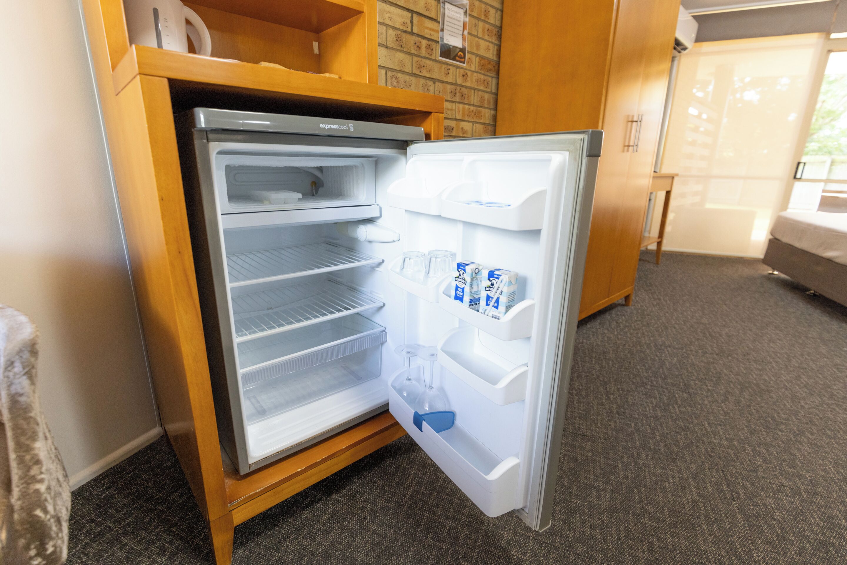 Un refrigerador compacto con varias estantes y un compartimento congelador, ubicado en una habitación de hotel con piso alfombrado y muebles de madera.