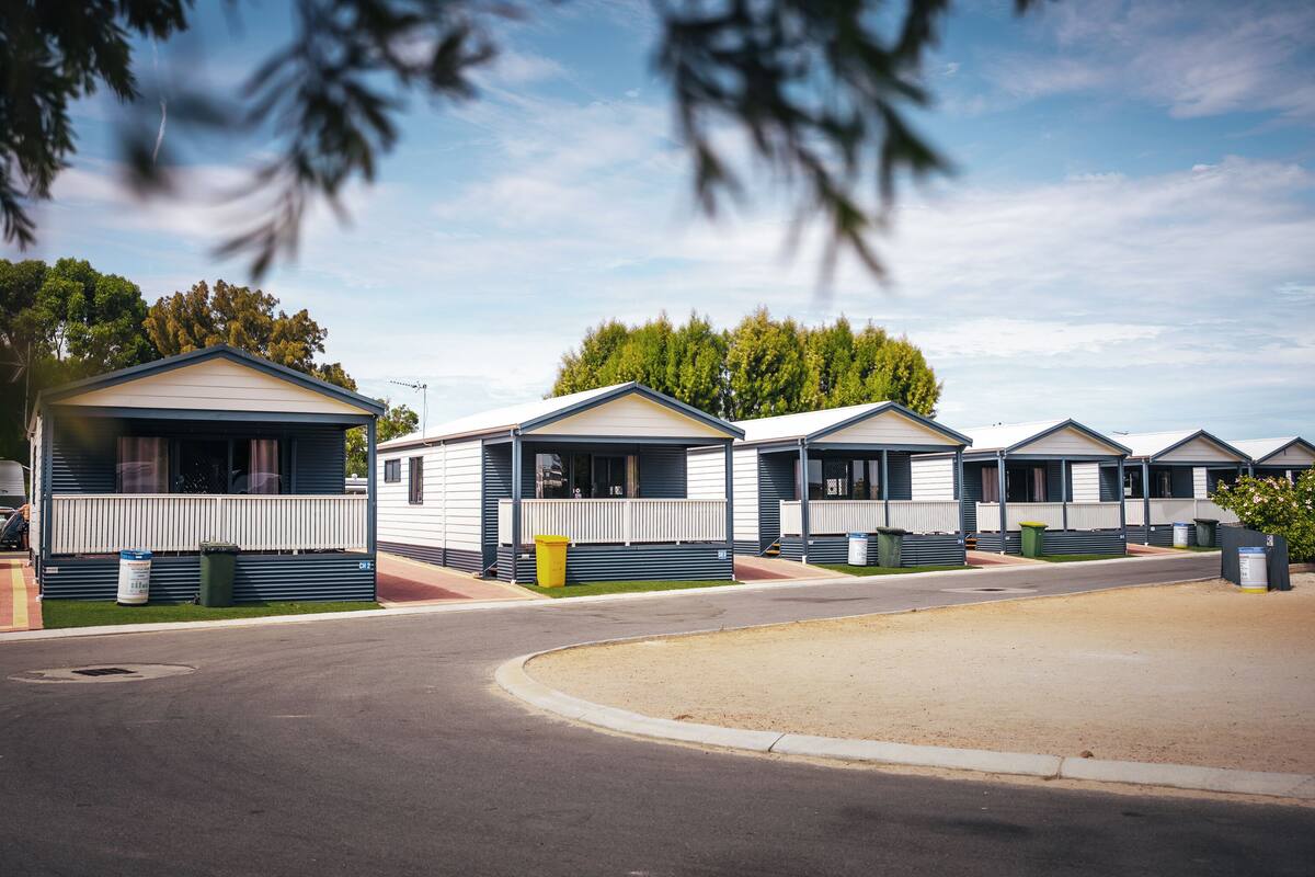 A row of mobile homes with a paved road in front, a grassy area, and trees in the background.