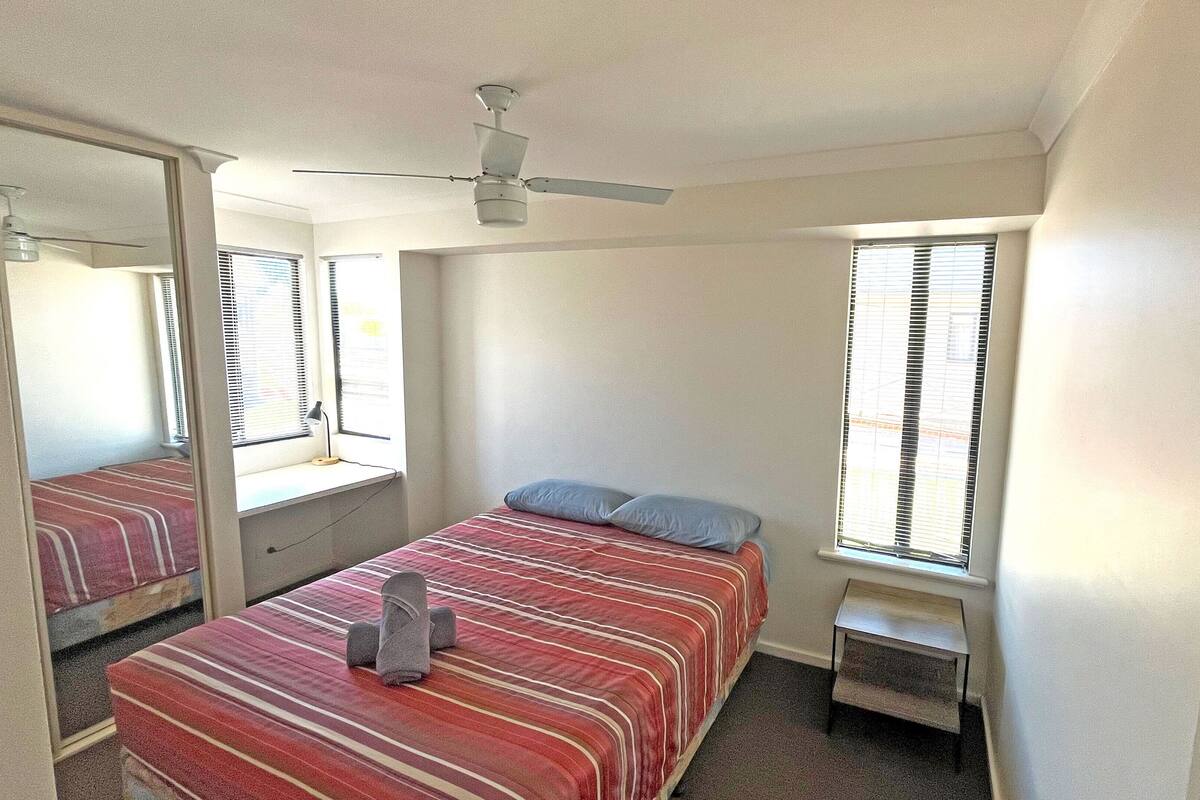 A bedroom with a bed, a small table, a chair, a ceiling fan, and a window with blinds.