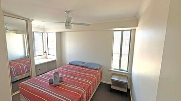 Un dormitorio con una cama, una mesita pequeña, una silla, un ventilador de techo y una ventana con persianas.