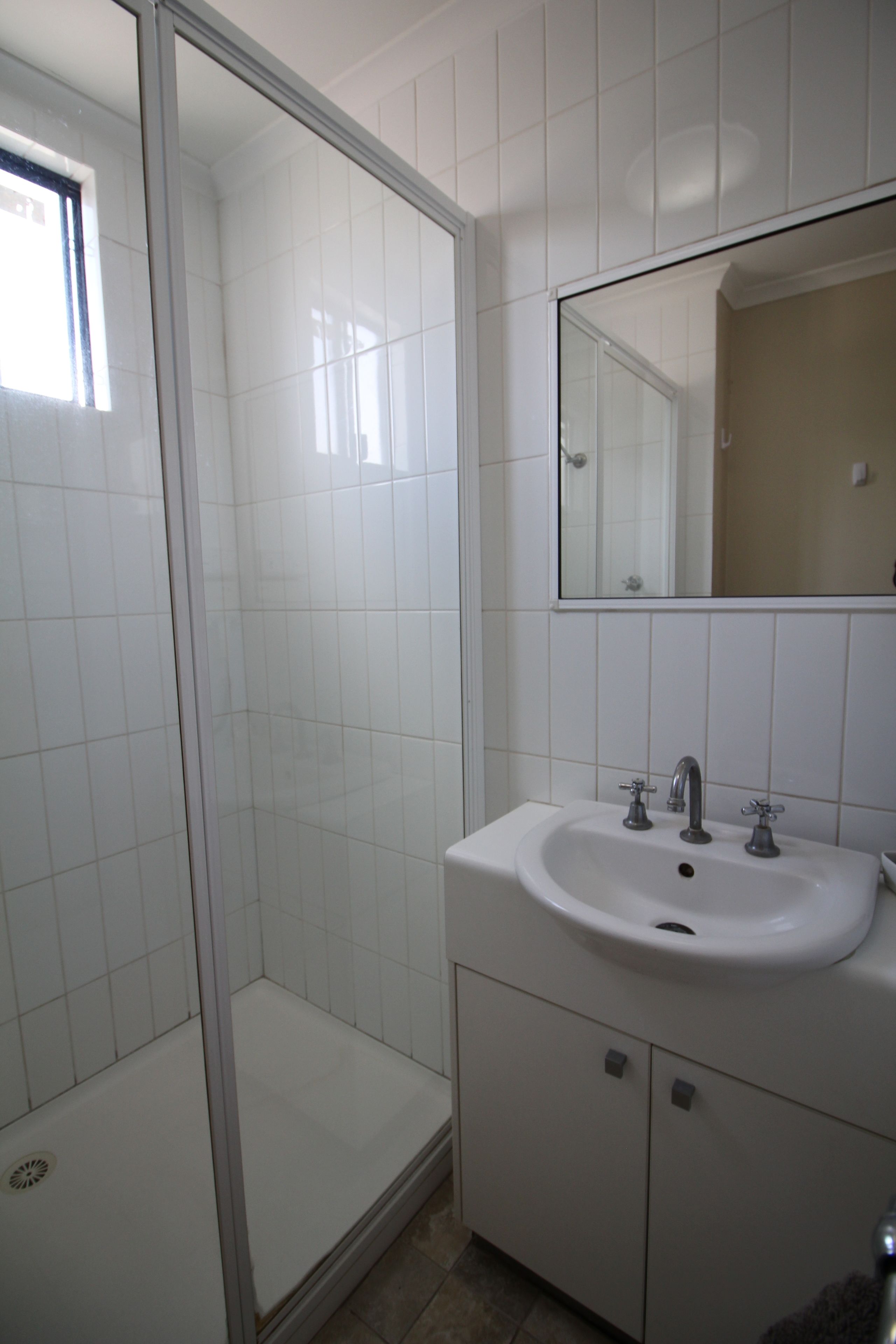 Ein Badezimmer mit weißen Fliesen, einer Glasdusche, einem Waschbecken mit zwei Wasserhähnen und einem Spiegel darüber.