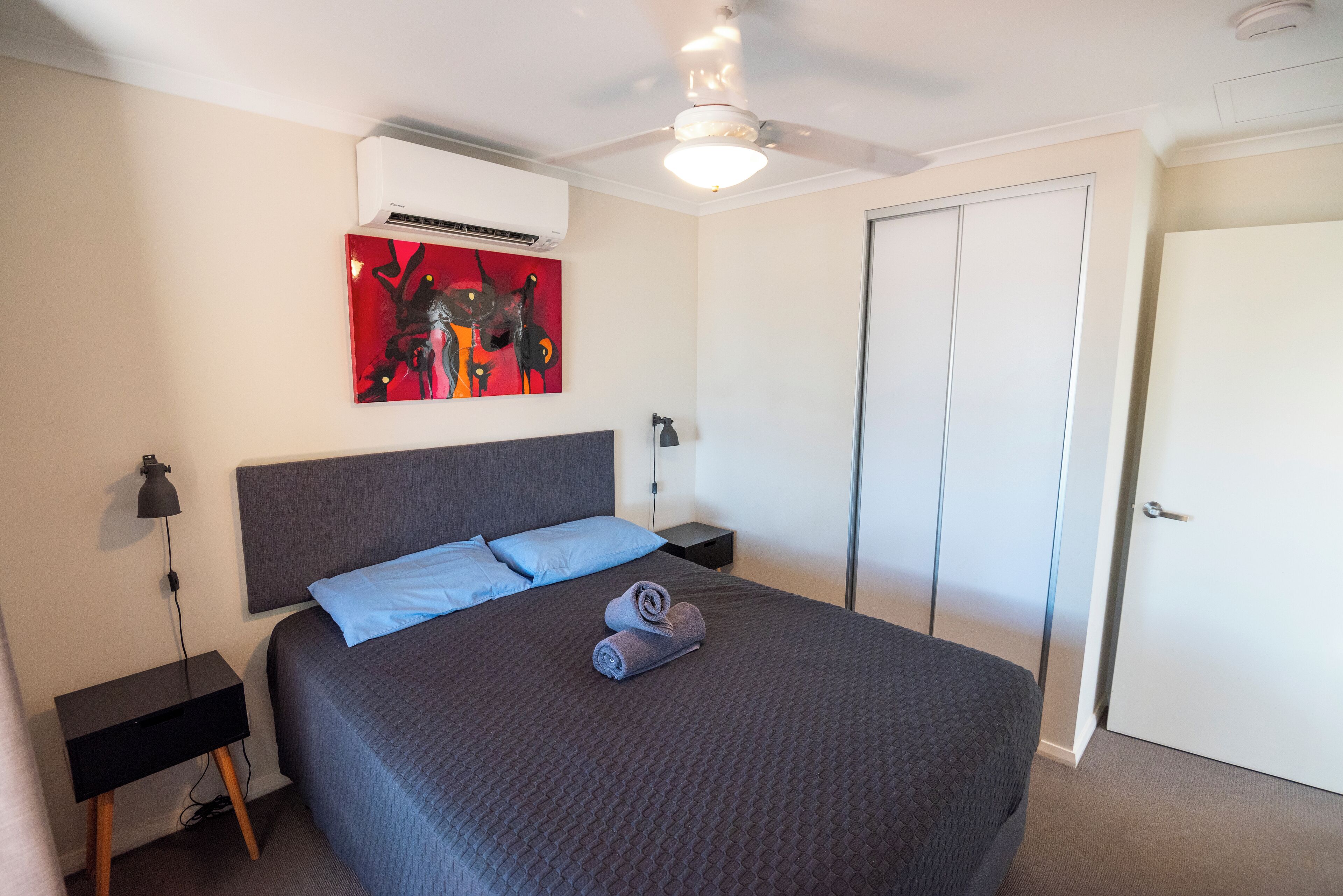 A neatly made bed with a dark quilt, two pillows, and rolled towels. There is a ceiling fan, a wall-mounted air conditioner, and a painting above the bed.