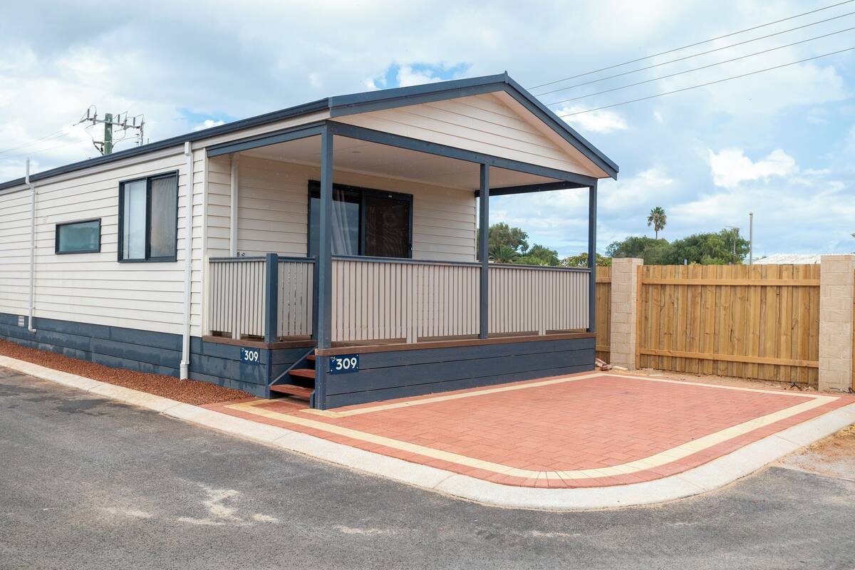 A single-story mobile home with a covered porch, a brick driveway, and a wooden fence.