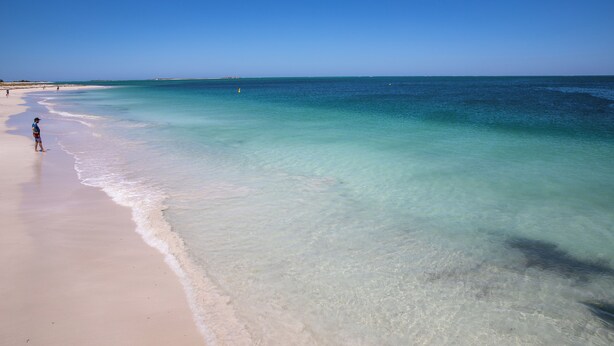 Une plage avec une eau turquoise et limpide, et une personne debout sur le sable.