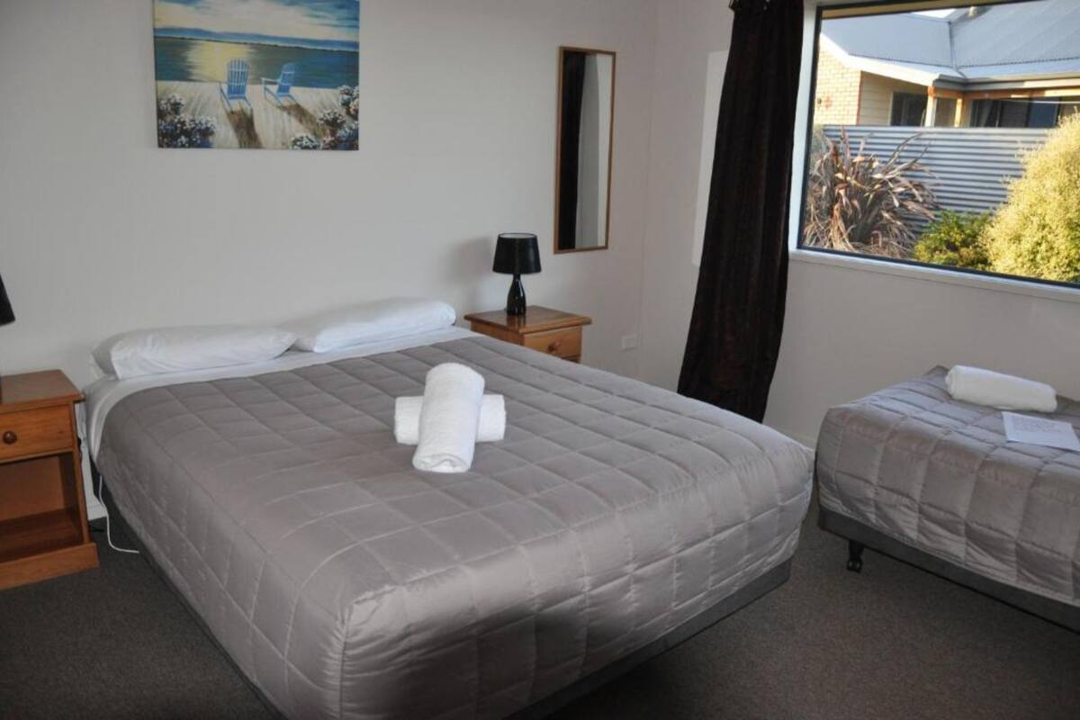 Un dormitorio con una cama grande, una cama más pequeña, una mesita de noche con lámpara, un cuadro en la pared y una ventana con vistas al exterior.
