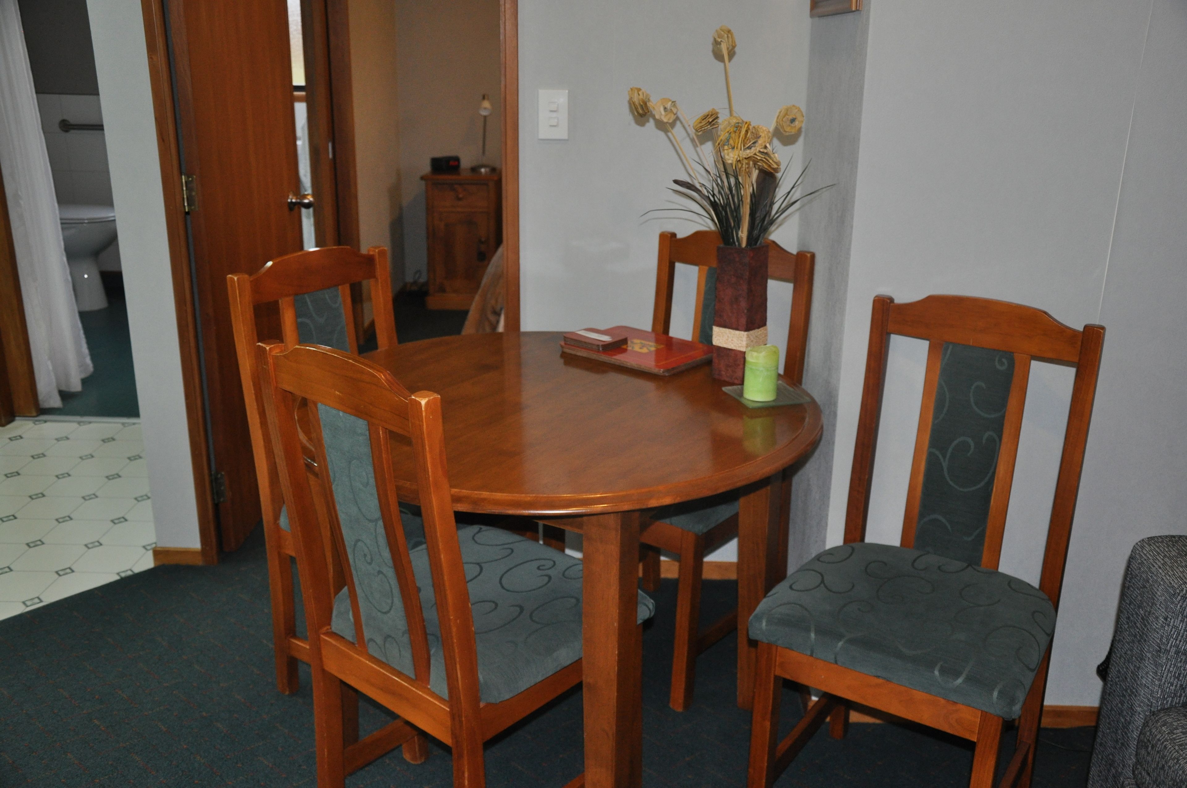 A wooden dining table with four chairs, a vase with dried flowers, and a small cabinet in the background.