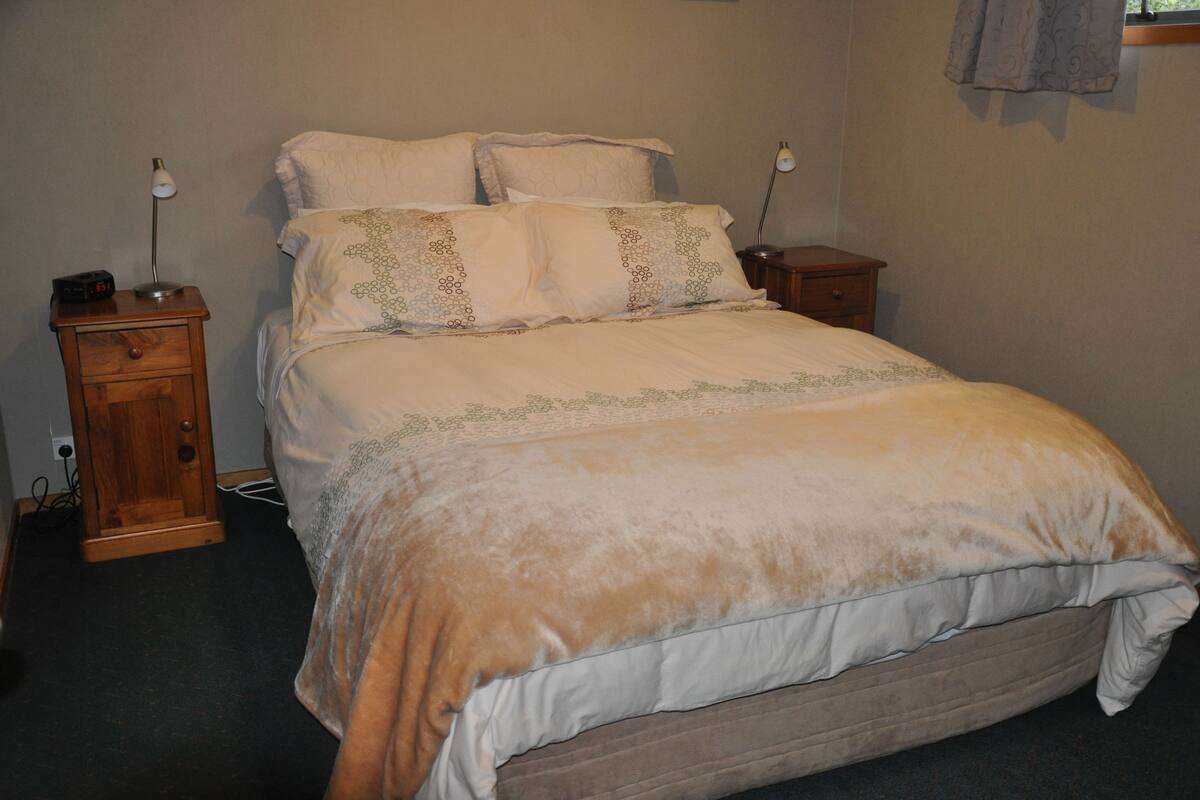 Una cama con edredón blanco y beige, dos mesitas de noche con lámparas y un mueble auxiliar de madera con cajón.