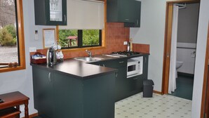 A compact kitchen with a central island, a built-in oven, a sink, and a window with blinds. - Birchwood Cottages (Te Anau)