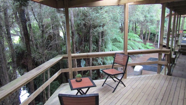 A wooden deck with two chairs and a small table, surrounded by dense forest. - The Crays Accommodation (Strahan)