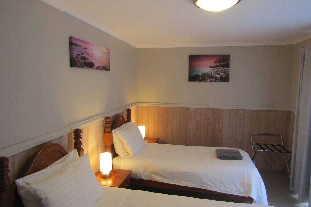 Habitación de hotel con dos camas individuales, cabeceros de madera, lámparas de noche y dos cuadros colgados en la pared.