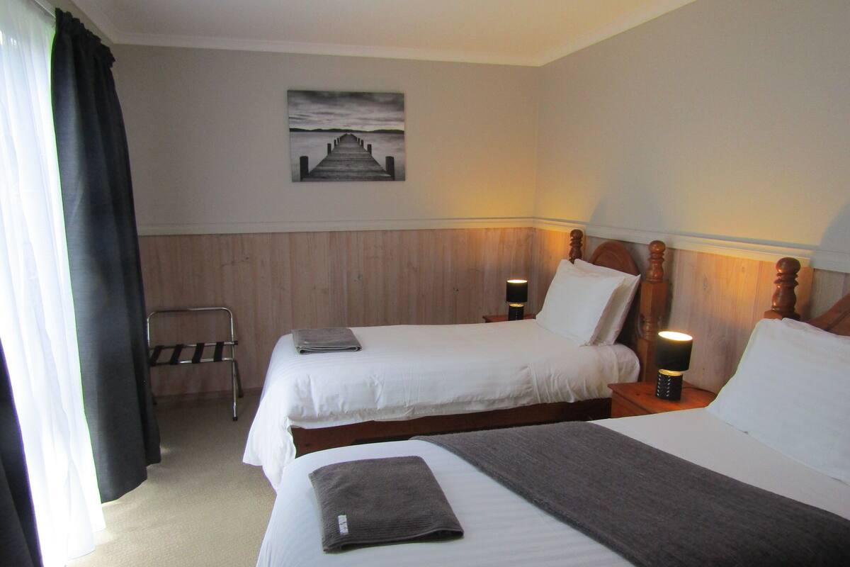 Habitación de hotel con dos camas, un cabecero de madera, una mesita de noche con lámpara, una silla y un cuadro en la pared.