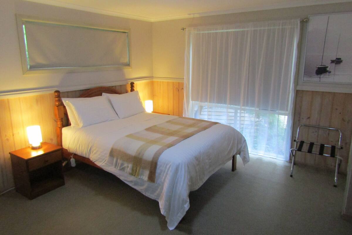 Una cama bien hecha con un cabecero de madera, dos lámparas de noche y una ventana con cortinas traslúcidas.