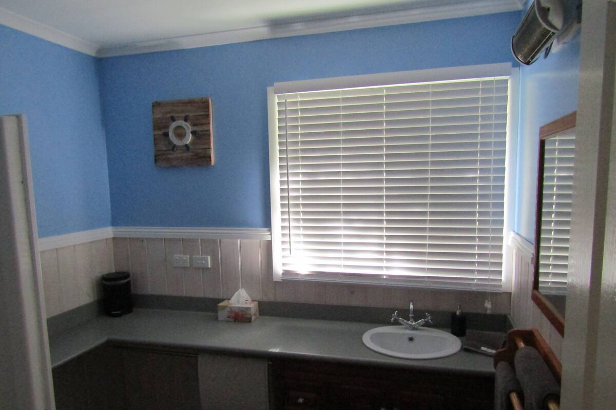 Un baño con lavabo, espejo, ventana con persianas y un adorno de pared de madera.