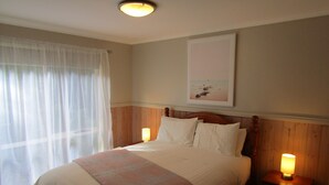 A neatly made bed with a headboard, two bedside lamps, a window with sheer curtains, and a framed picture on the wall. - The Crays Accommodation (Strahan)