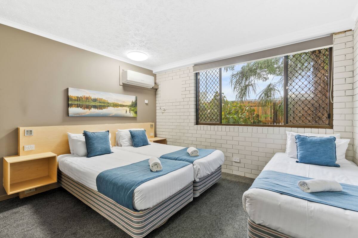 A hotel room with two beds, a large window with a view, a painting on the wall, and a small shelf.