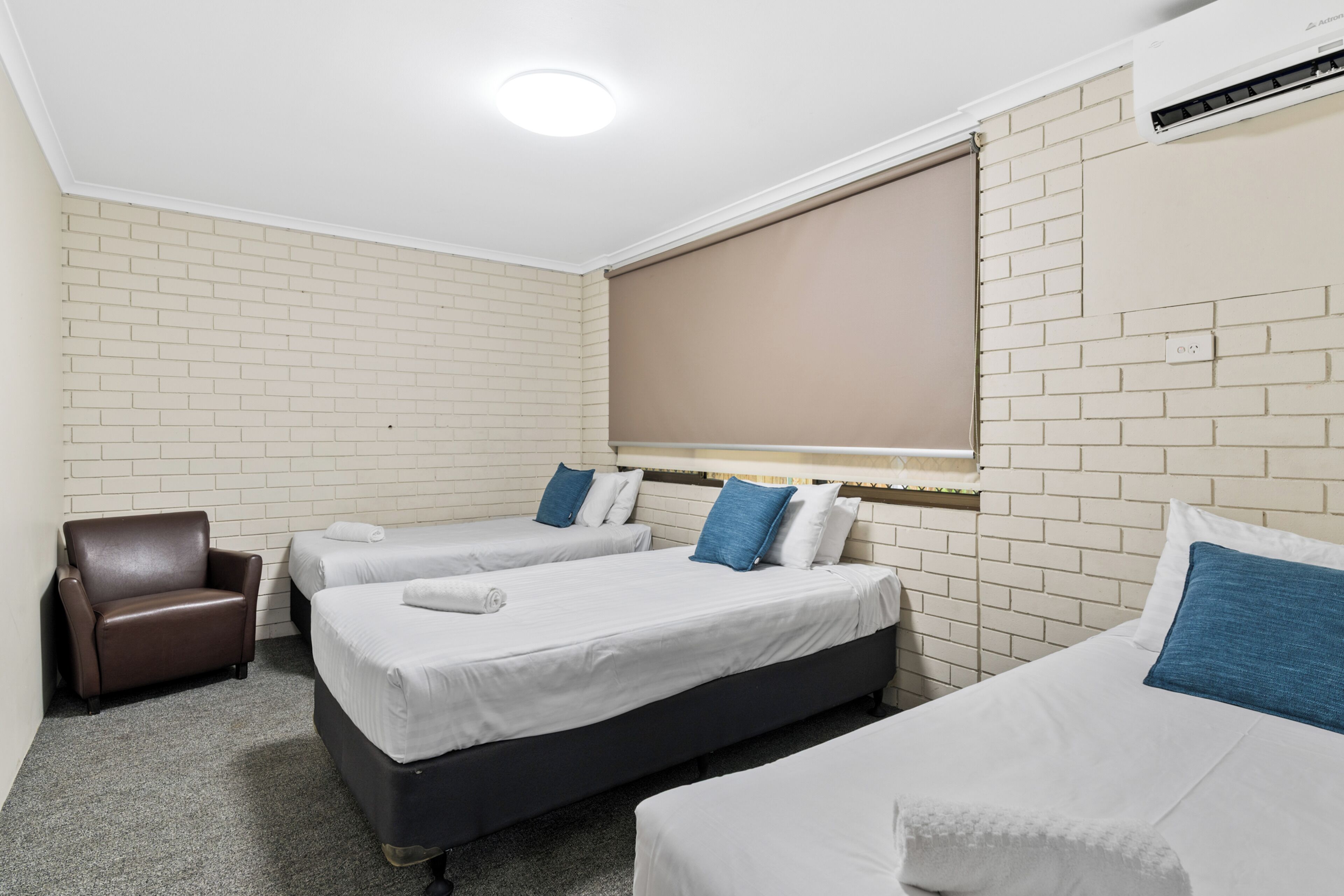 A hotel room with two beds, a brown armchair, and a window with blinds.