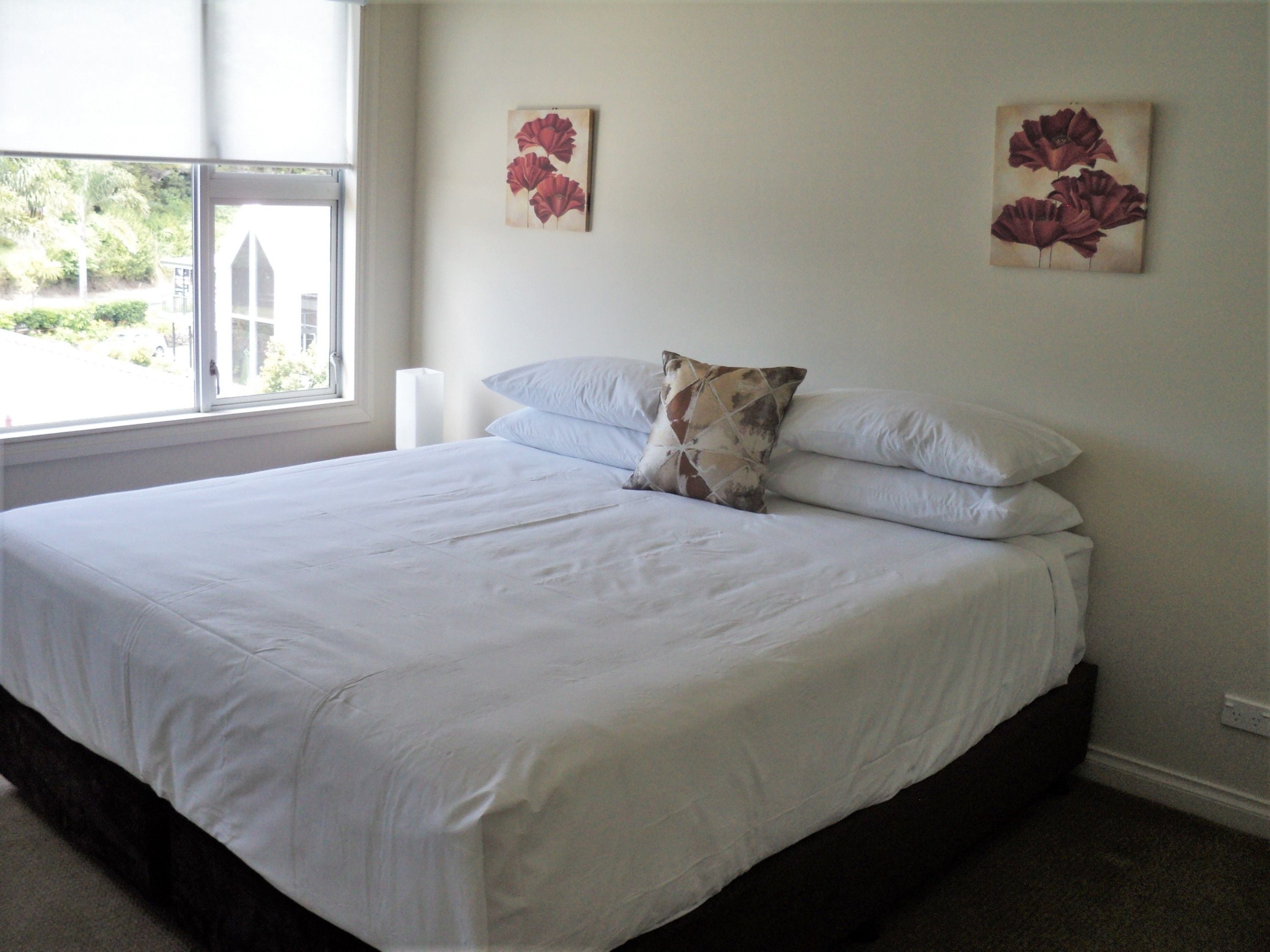 A neatly made bed with white linens and a single decorative pillow, a window with a view of greenery, and two wall-mounted floral paintings.