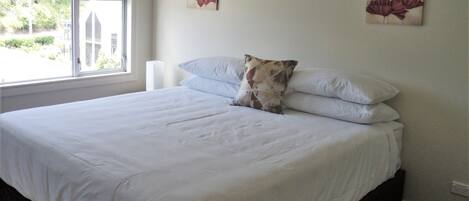 A neatly made bed with white linens and a single decorative pillow, a window with a view of greenery, and two wall-mounted floral paintings.