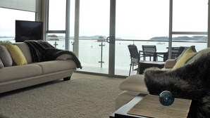 A modern living room with a sofa and armchair facing a large window overlooking a body of water. - Edgewater Palms Apartments (Paihia)